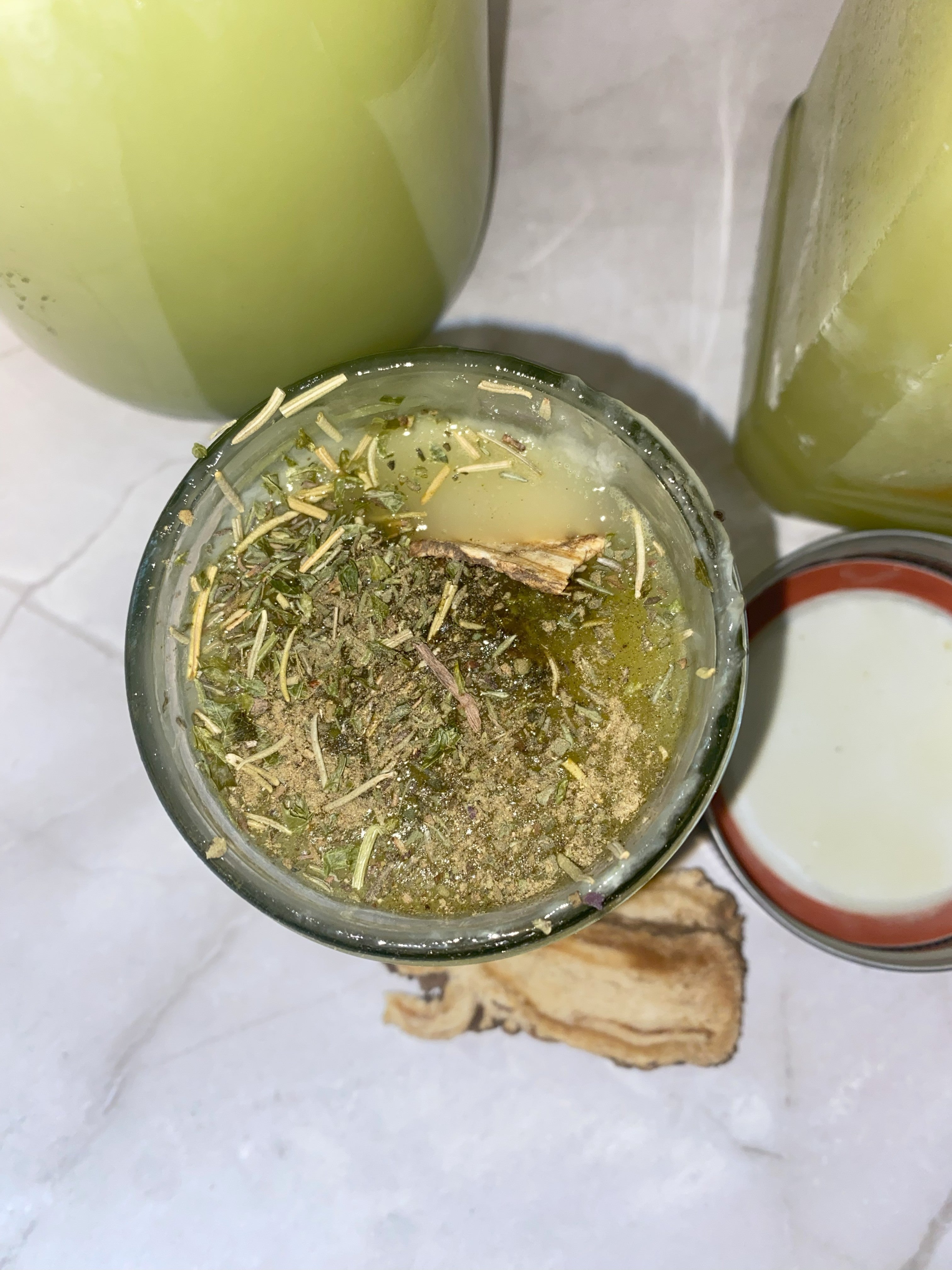 Open jar of green herbal mixture with a small glass jar container of cleansing ritual bath on a white surface.