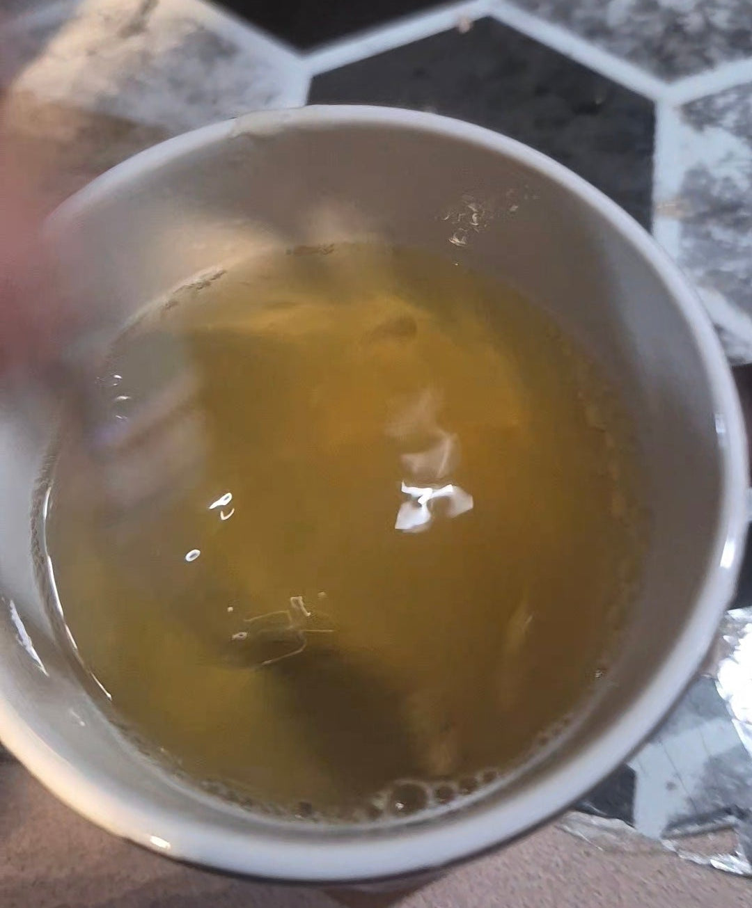 A close-up of a white cup containing a mushroom herbal and root tea, with a hand holding a spoon near the rim. The background shows a gray and black hexagonal tile floor.